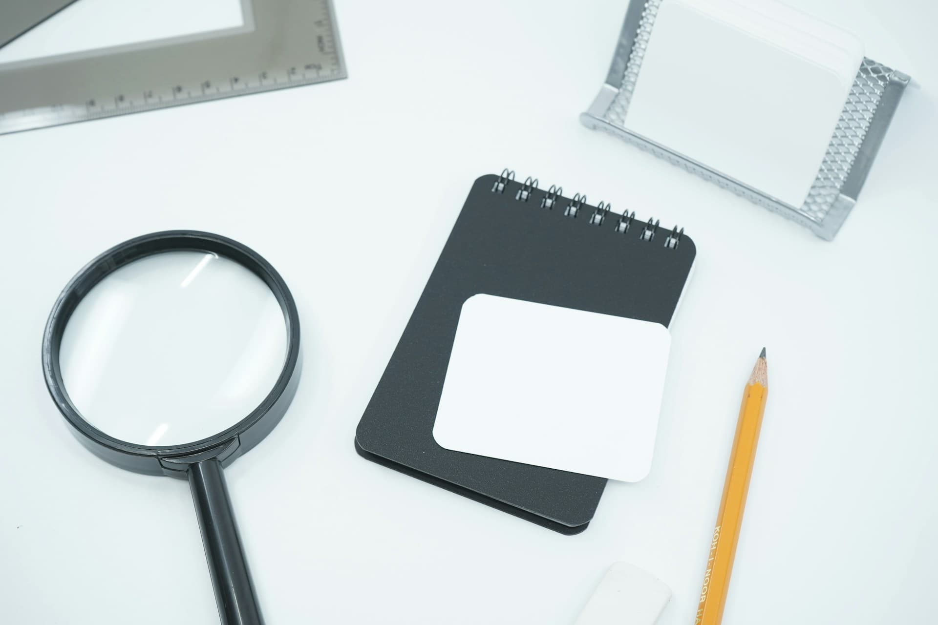 Magnifying glass, notepad, pencil and ruler arranged on a white desk with a blank card