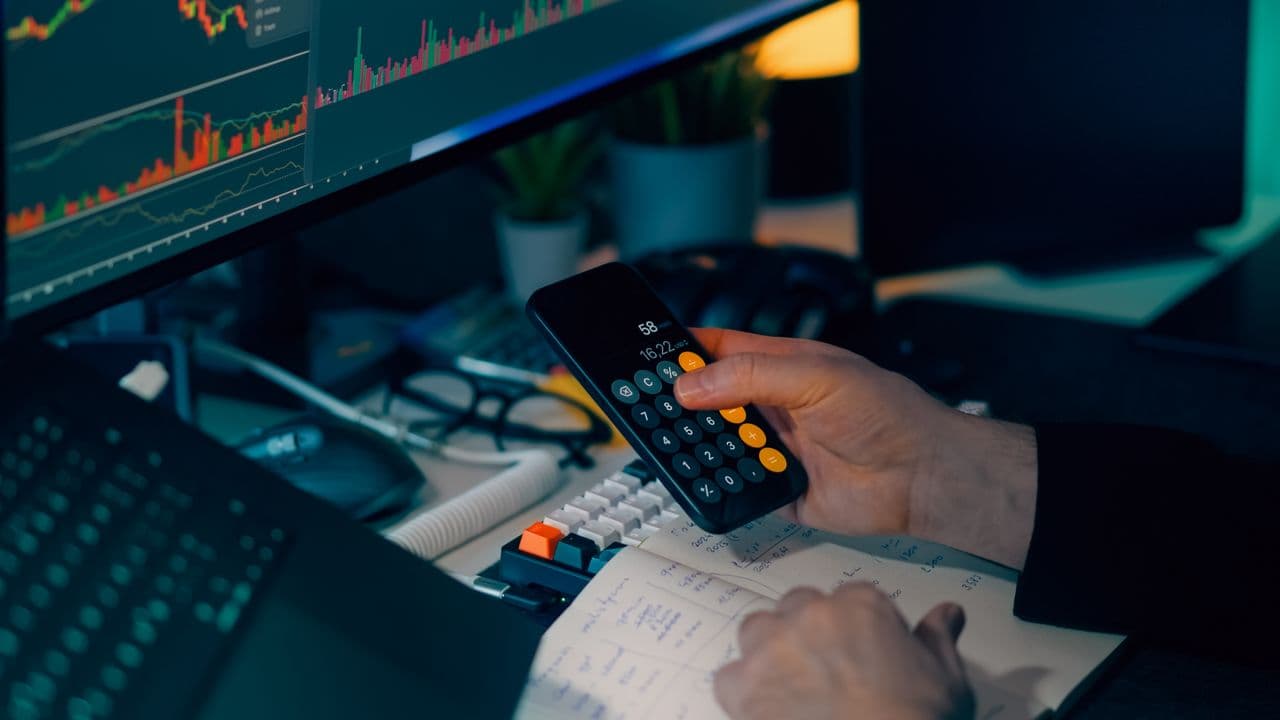 Trader using a phone calculator at a desk with handwritten notes and candlestick charts on screen