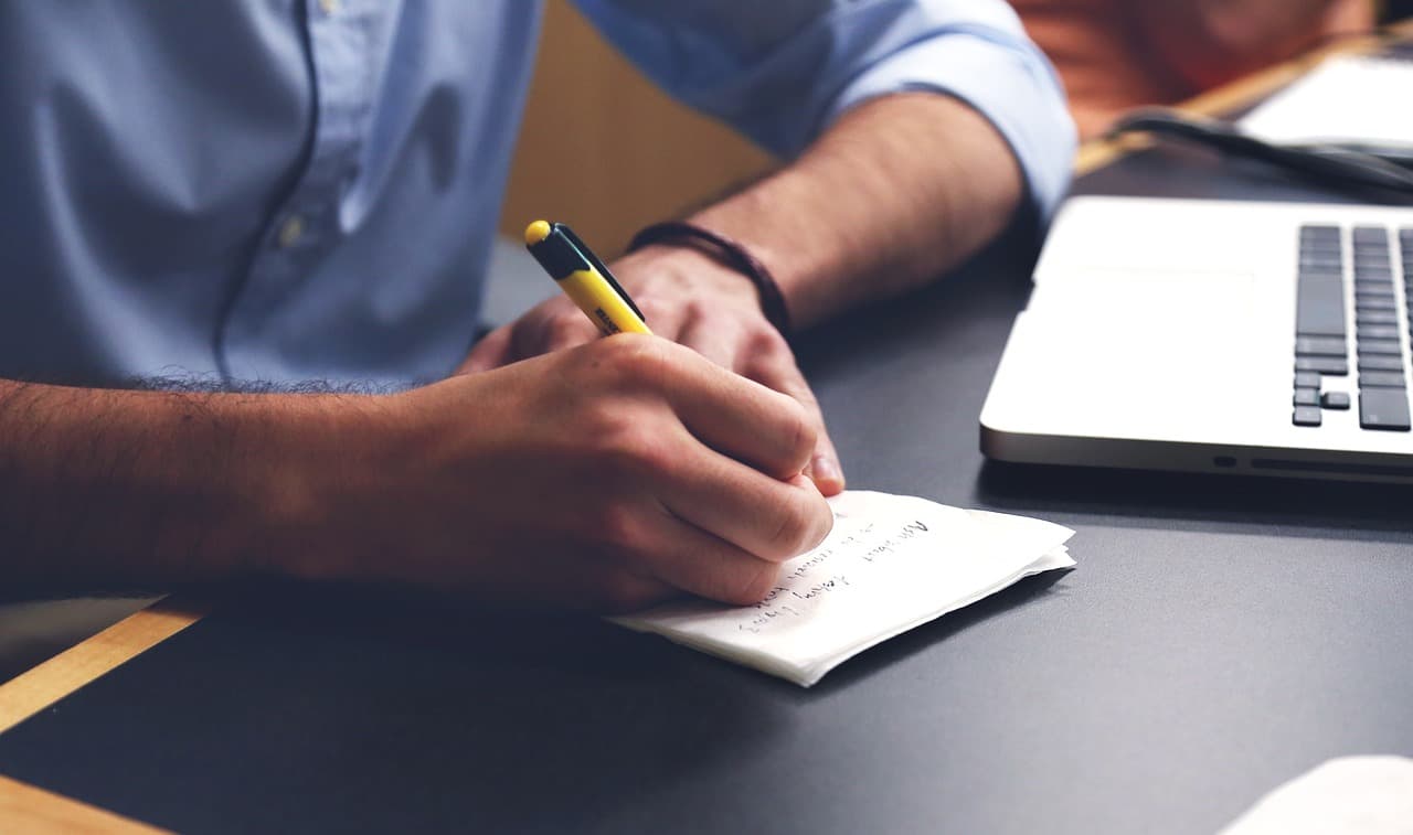 Person writing notes on a folded sheet of paper at a desk beside an open laptop