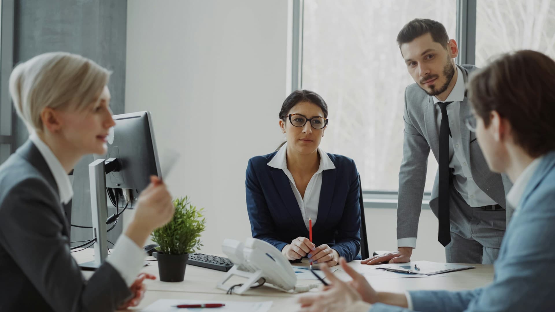 Four colleagues in business suits discussing paperwork around a desk in a bright office